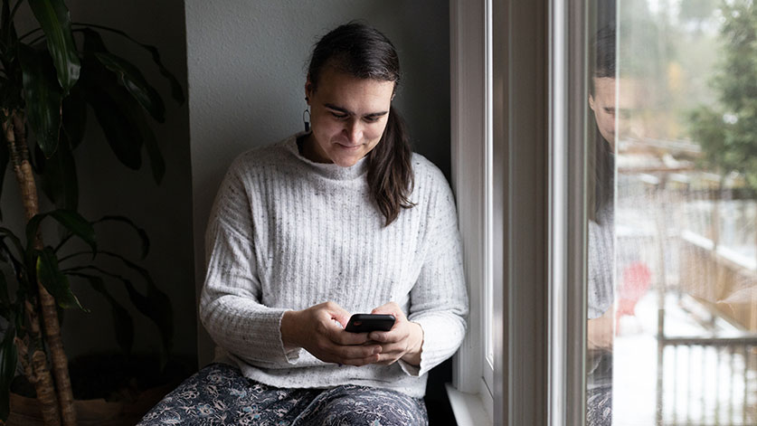 weibliche Person sitzt am Fenster und sucht zuversichtlich in ihrem Handy nach Wege aus der Arbeitslosigkeit