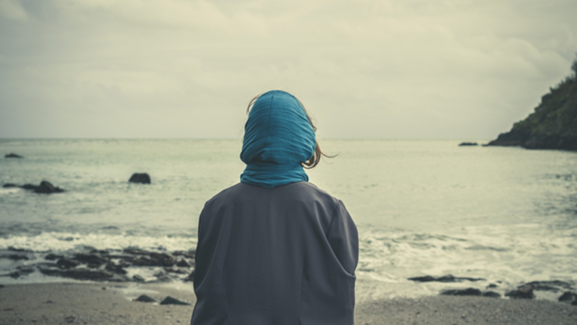 Person* mit blauem Tuch um den Kopf und grauer Kleidung steht mit dem Rücken zur Kamera am Strand und blickt aufs Meer. Der Himmel ist bewölkt, die Stimmung wirkt ruhig und nachdenklich.