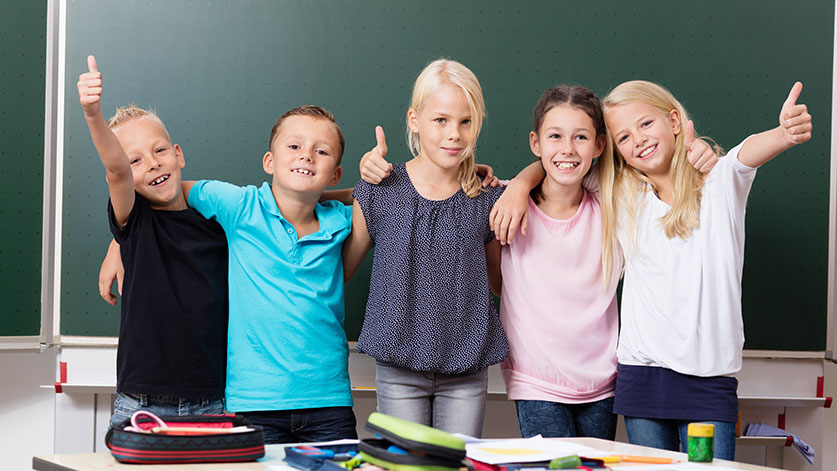 Fünf Kinder stehen Arm in Arm vor einer grünen Tafel im Klassenraum. Sie lächeln in die Kamera und zeigen Daumen hoch. Auf dem Tisch davor liegen bunte Schulmaterialien.