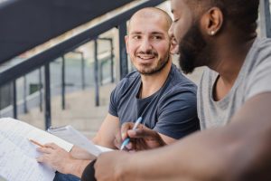 Zwei junge Männer sitzen draußen auf einer Treppe und lernen gemeinsam, während sie Notizen und Unterlagen durchgehen. Sie unterhalten sich freundlich, was eine positive Atmosphäre vermittelt.