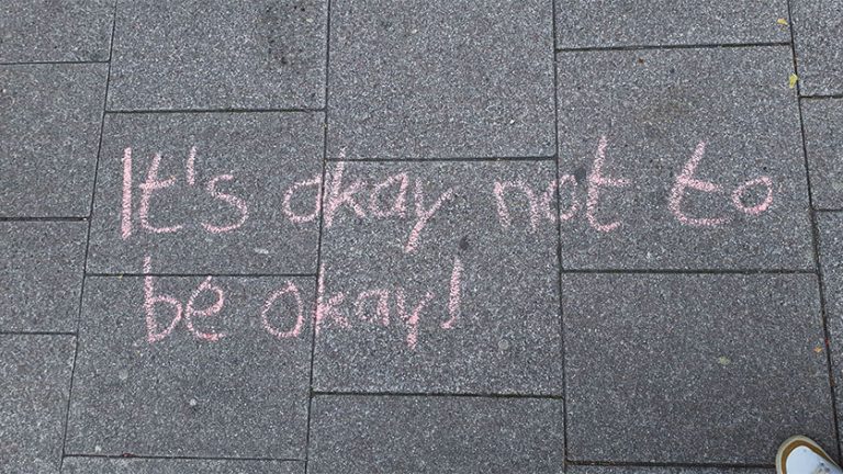 Mit rosa Kreide steht auf grauen Pflastersteinen geschrieben: „It's okay not to be okay!“ – Eine positive Botschaft zur mentalen Gesundheit.