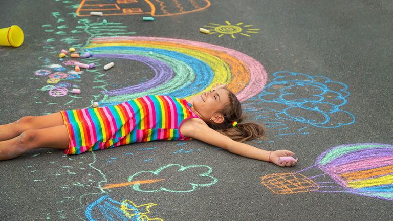 Ein Mädchen in einem bunten Regenbogenkleid liegt lachend auf einer Straße, die mit farbenfroher Kreide bemalt ist. Zu sehen sind ein Regenbogen, eine Sonne, ein Heißluftballon, Wolken, Bäume und ein Haus. Die Szene wirkt fantasievoll und verspielt.