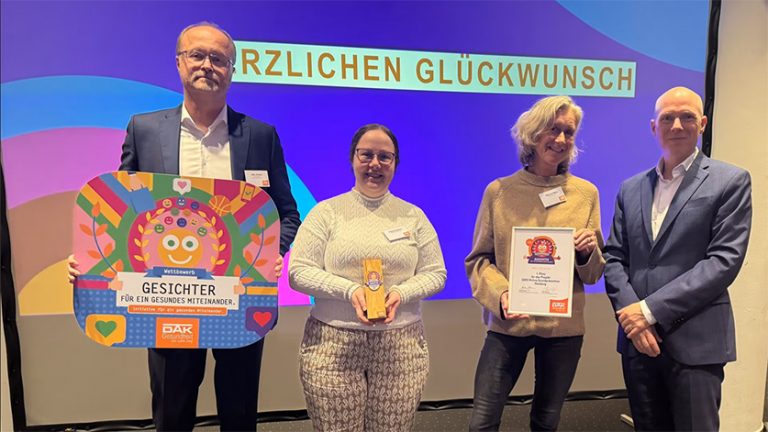 Jens Juncker, Paula Koudmani, Nina von Ohlen und Tim Angerer stehen nebeneinander vor einer Leinwand mit der Aufschrift „HERZLICHEN GLÜCKWUNSCH“. Es handelt sich um die Preisverleihung „Gesichter für ein gesundes Miteinander“ von der DAK-Gesundheit für das Projekt [U25] Hamburg von IN VIA Hamburg.