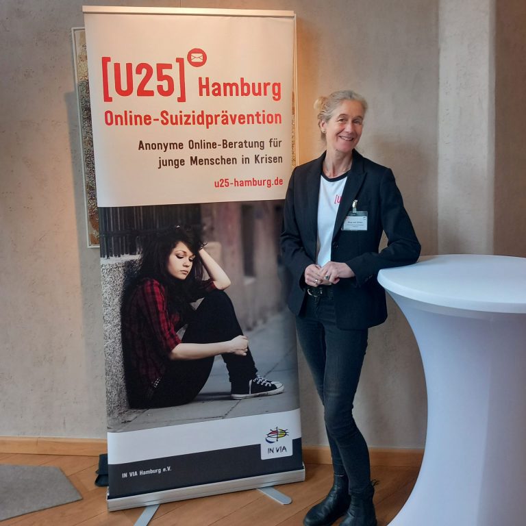 Nina von Ohlen, Vorstandsmitglied der Deutschen Gesellschaft für Suizidprävention (DGS), steht lächelnd neben einem Roll-Up von [U25] Hamburg. Das Banner informiert über die anonyme Online-Beratung für junge Menschen in Krisen zur Suizidprävention. Darauf zu sehen ist auch ein Bild eines nachdenklich sitzenden jungen Mädchens. Das Angebot wird von IN VIA Hamburg e.V. getragen.